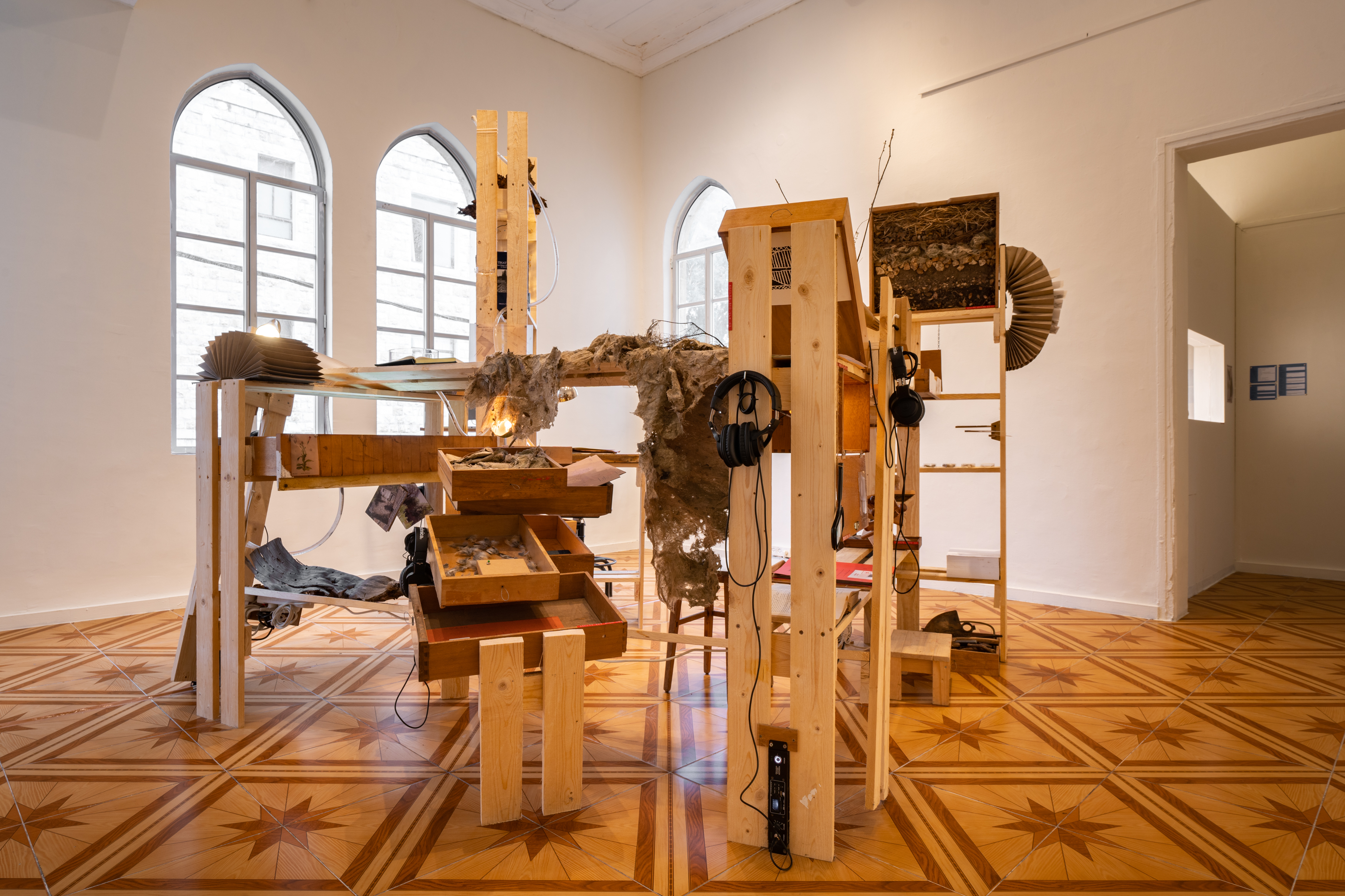 Sculpture installation by Maayan Tsadka and collaborators Nawal Arafat and Leoni Schein in a gallery space.  The installation consists of a series of constructed wooden architectural forms reminiscent of shelves, drawers,  ladders, and other home objects. There are small piles of organic materials, archival materials, photographs,  cups, rocks, books, pvc tubing, an opened accordion folder, among other items throughout. There are also  headphones rigged to electrical interfaces present throughout the installation. The floor within the gallery  space is ornamental and tiled with repeated star-like forms.