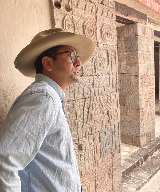 Portrait of Edgar Garcia in Teotihuacan, an ancient Mesoamerican city in Mexico. Garcia is wearing a faded light blue button down shirt, a tan wide brimmed hat, and tortoise eyeglasses. In the background is a row of rectangular stone pillars carved with what appears to be glyphs and symbols representing the cosmos and nature. Garcia is captured in quarter profile looking outwards.