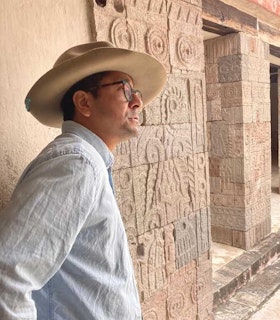 Portrait of Edgar Garcia in Teotihuacan, an ancient Mesoamerican city in Mexico. Garcia is wearing a faded light blue button down shirt, a tan wide brimmed hat, and tortoise eyeglasses. In the background is a row of rectangular stone pillars carved with what appears to be glyphs and symbols representing the cosmos and nature. Garcia is captured in quarter profile looking outwards.