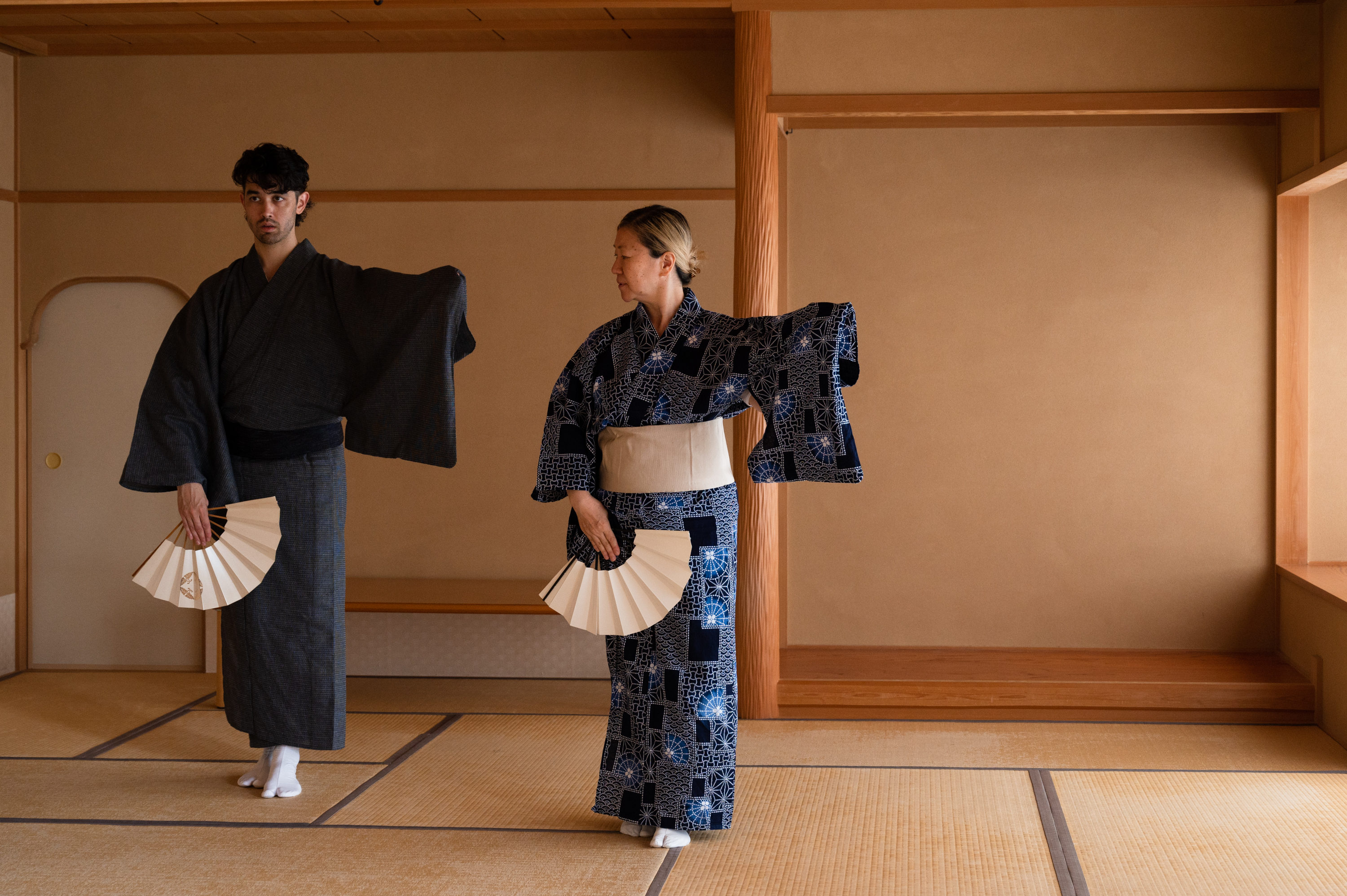 Benjamin Akio Kimitch and Yasuko Yokoshi dancing in Kyoto, Japan. Kimitch  is seen mirroring Yokoshi’s movement, and each has one foot ahead of the  other and their lefts arm out to the side. Kimitch and Yokoshi eachwear  kimonos, obi belts, and tabi socks and are holding a sensu fan in their right hand.