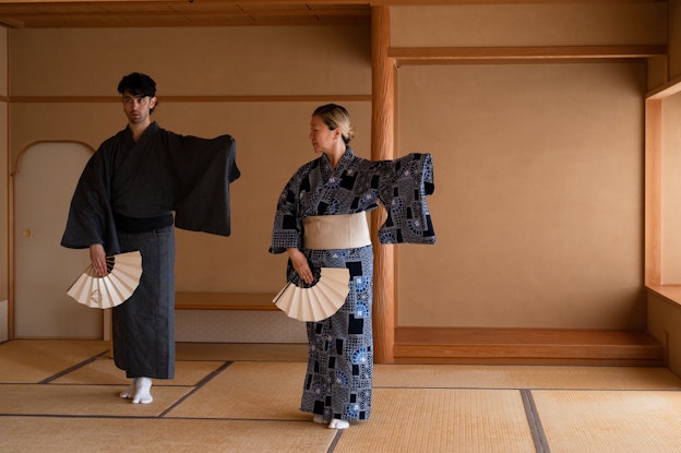 Benjamin Akio Kimitch and Yasuko Yokoshi dancing in Kyoto, Japan. Kimitch  is seen mirroring Yokoshi’s movement, and each has one foot ahead of the  other and their lefts arm out to the side. Kimitch and Yokoshi eachwear  kimonos, obi belts, and tabi socks and are holding a sensu fan in their right hand.