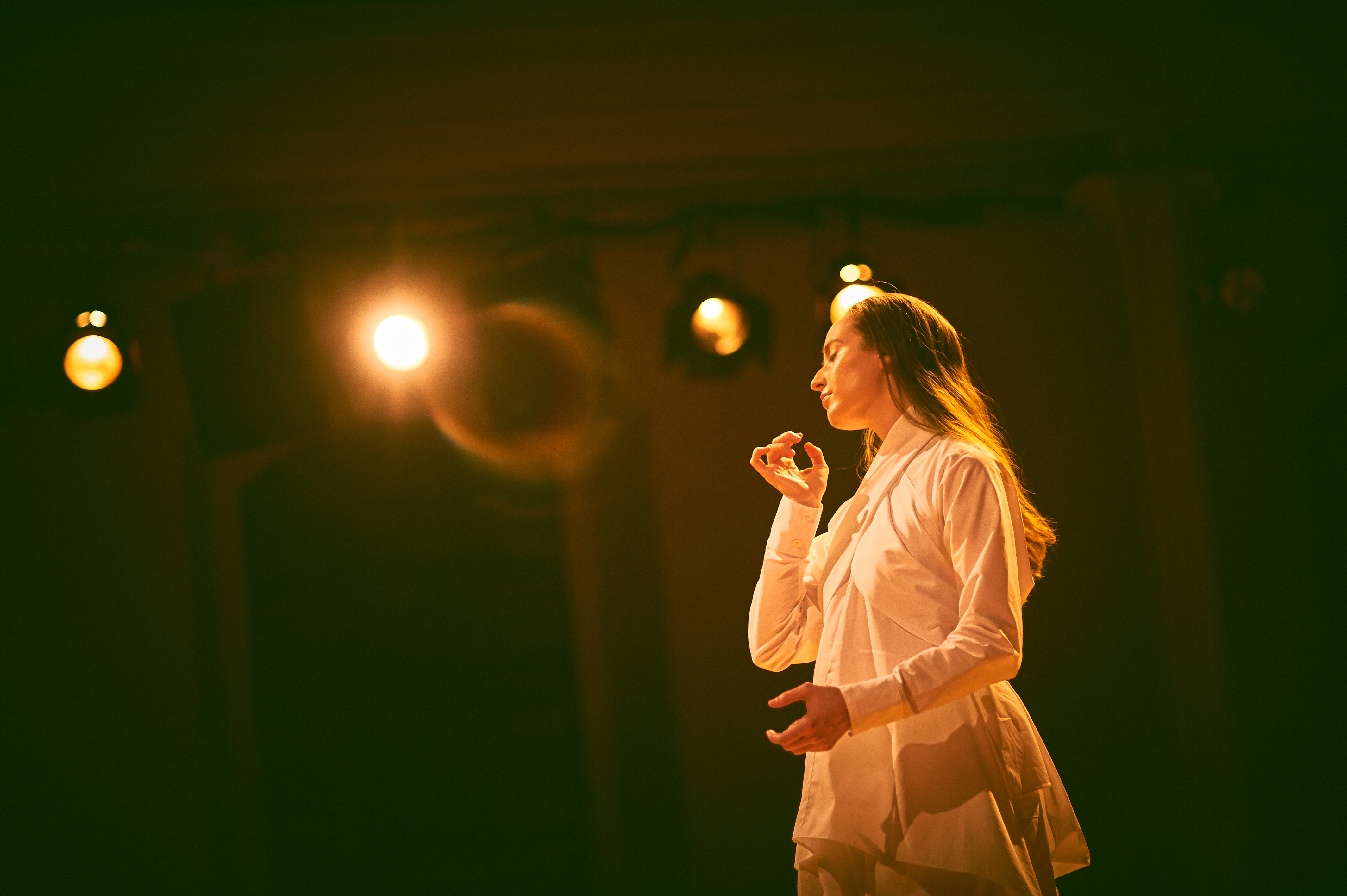Julie McMillan Castellano dancing at The Noguchi Museum in a white  long sleeve wrapped shirt. They are looking down towards the ground  as one hand is by their shoulder and the other is in front of their stomach. Both of their hands are formed in the shape of “C”. The stage lighting basks  Julie in a golden glow, while the image itself has a green fog.