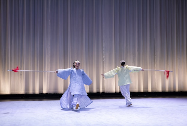 Lai Yi Ohlsen and Pareena Lim performing, moving in a circle.  They are both wearing costumes inspired by Eastern Asian performance  traditions. Lai Yi Ohlsen is facing the audience is in light purple dress, while  Pareena Lim, facing back stage, is in light yellow. Both of their hands are in  the air to their sides, and they each have one foot in front of the other.  Behind the duo is a white curtain that is just a bit short of reaching the  ground, the pitch-black of back stage is visible from below.  The floor is light purple, matching Lai’s dress.