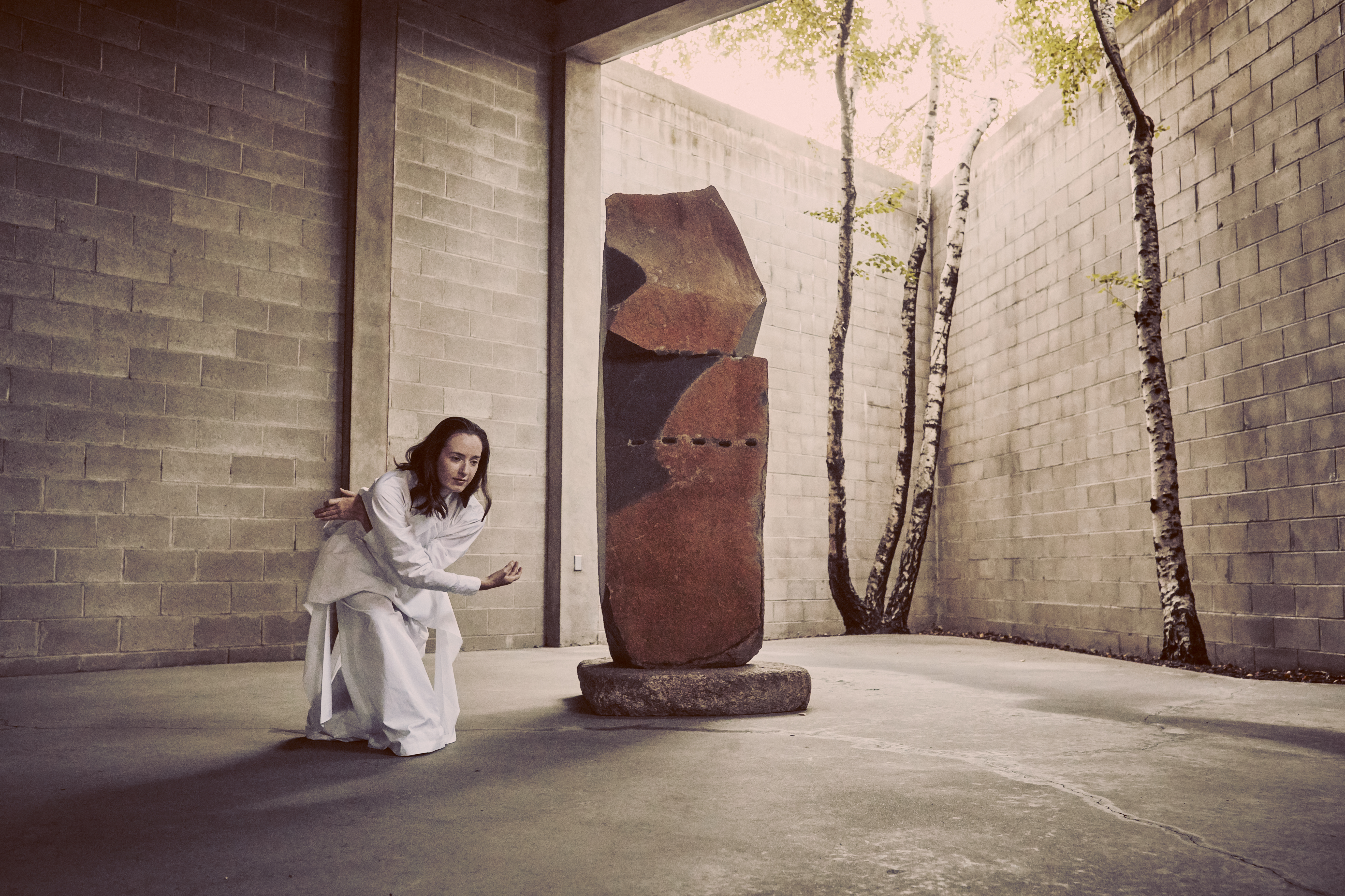 Julie McMillan Castellano dancing in the indoor/outdoor gallery of  The Noguchi Museum. In a white long sleeve shirt and white pants,  Julie’s knees are bent and their arms are interlacing one another. To  Julie’s side is a Noguchi sculpture. In the upper right hand corner, the  sun is shining onto the concrete block wall, two paper birches, and  the pavement.