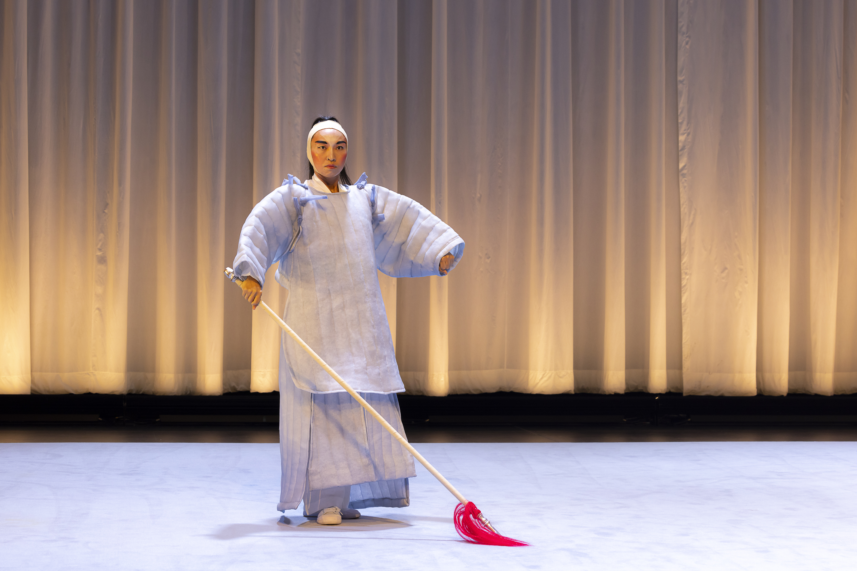Lai Yi Ohlsen is wearing a light purple costume inspired by Eastern Asian  performance traditions. Their hands are to their sides and they are making  fists with their hands. Behind Lai is a white curtain, glowing with yellow  lighting. Matching their dress, the floor is also light purple.