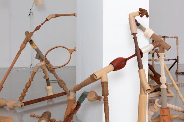 Close up view of “Negotiated Difference,” a rhizomatic sculpture made out of carved wood and  3D-printed filaments in wood, metal, and plastic, connected with various objects including  balusters, pipes, and a tennis racket without strings. The sculpture is installed on a gallery  floor, wrapping around a very tall white pedestal.