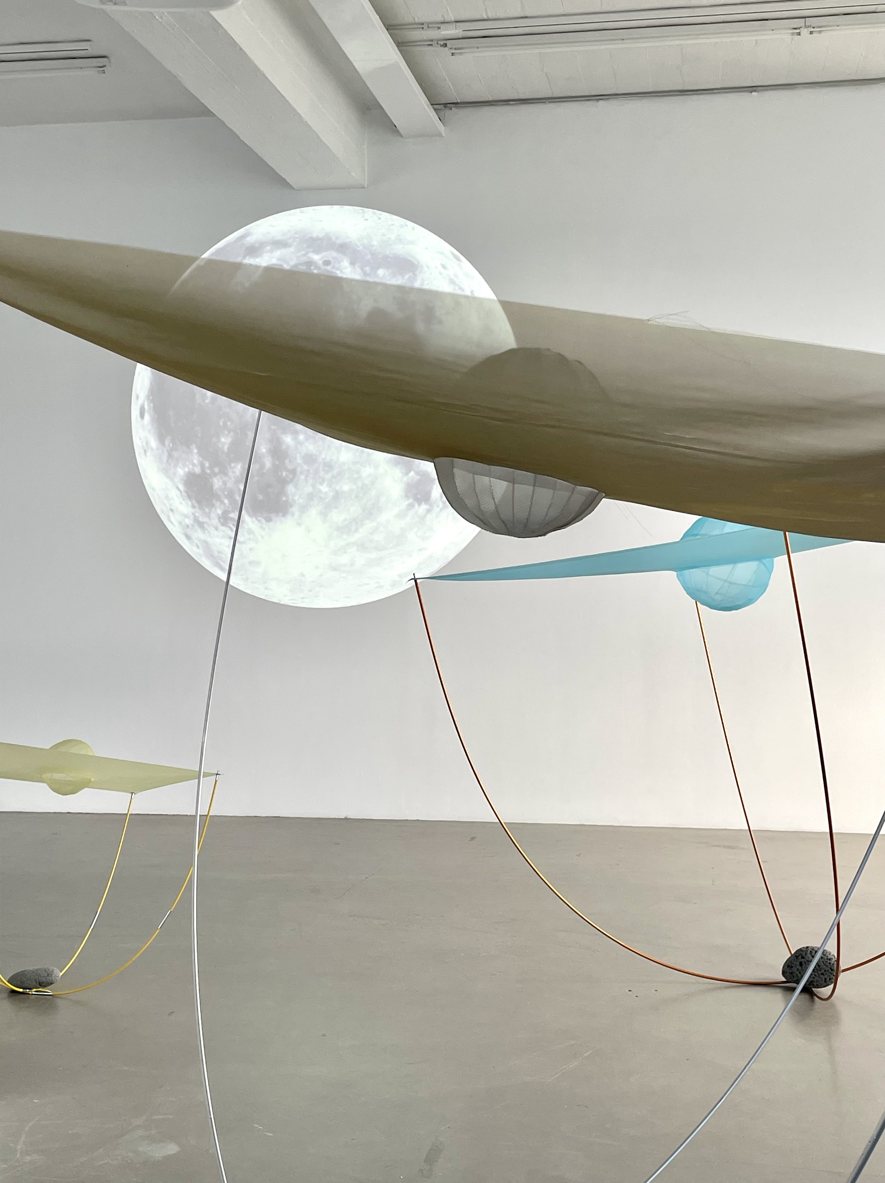 Installation view of “Bobbing Moons.” A full moon is projected onto a white gallery wall with  three structures that resemble tidal turbine systems, one beige and white, another green,  and one blue, orange, and yellow, all of which are balanced on the light grey cement floor.