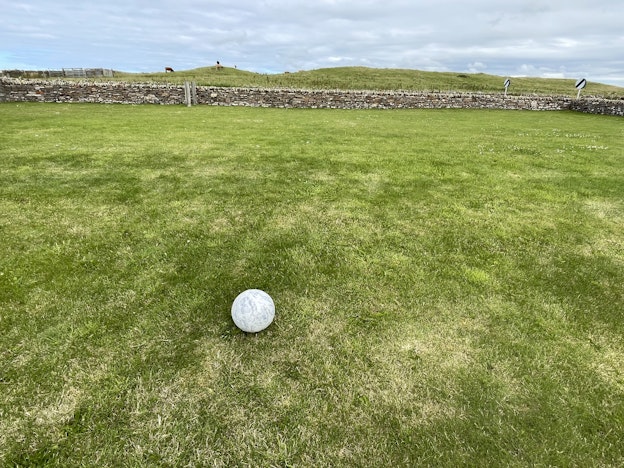 “Orkney Moon,” a print of the moon, is installed on a flat grassy field during a partly cloudy  day. The moon itself is very small compared to the field. In the distance, a horizontal stone  wall borders the flat field from hills, which have cattle grazing throughout. There is also a  pen for the cattle in the background.
