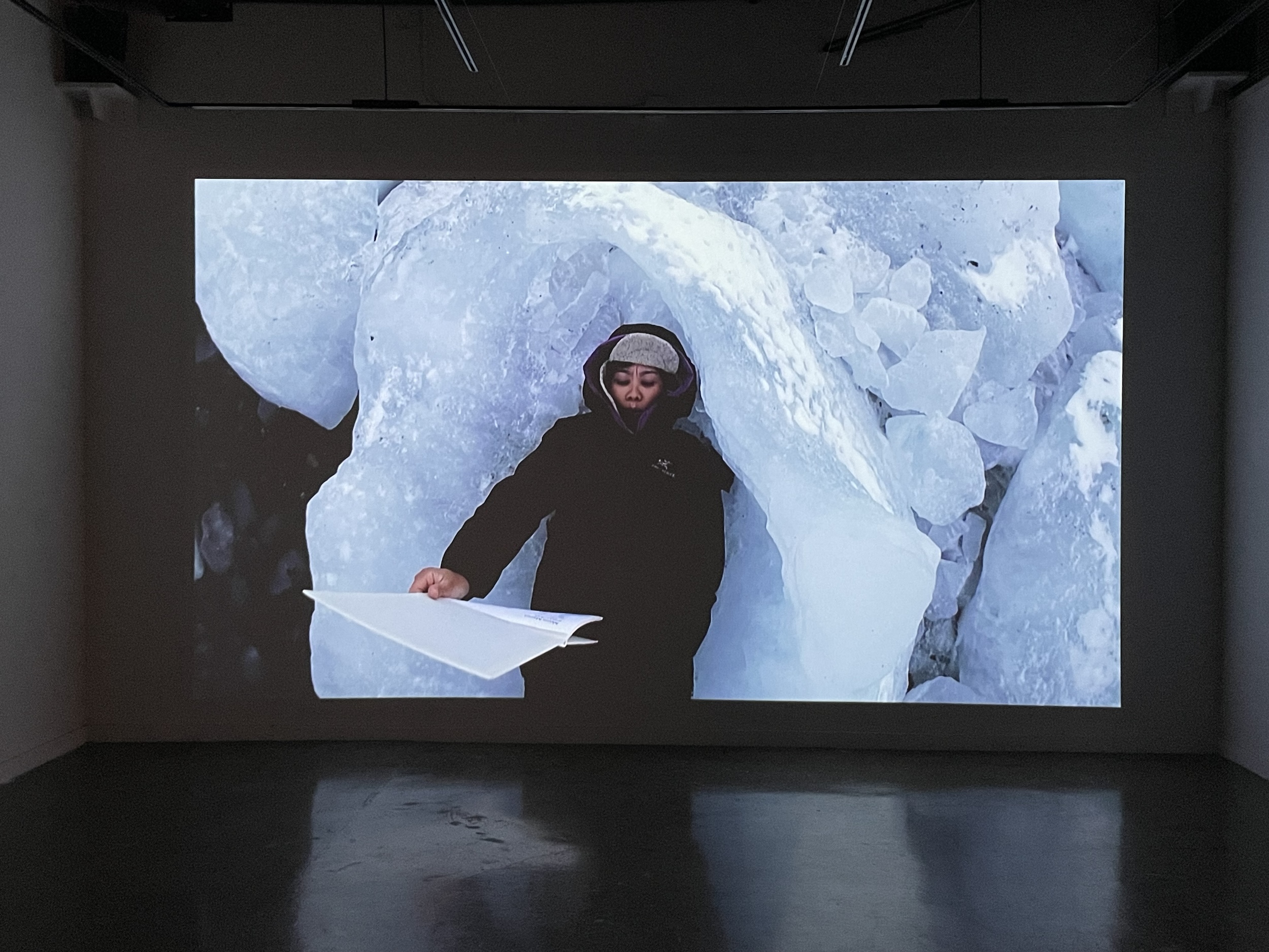 Installation view of “The Arctic Circle,” a video projection on a white gallery wall in a dimly lit  room. The still is of a person who appears to be lying down and reading on a big chunk of  ice in a tundra. Their hand is extended in front of them, holding a large booklet printed onto  white paper. They are wearing a black winter jacket, which is zipped up over their mouth, a  white cap with grey specks that almost matches the ice, and the hood from their jacket,  which is accented with purple around its edges. The projection reflects onto the cement floor.