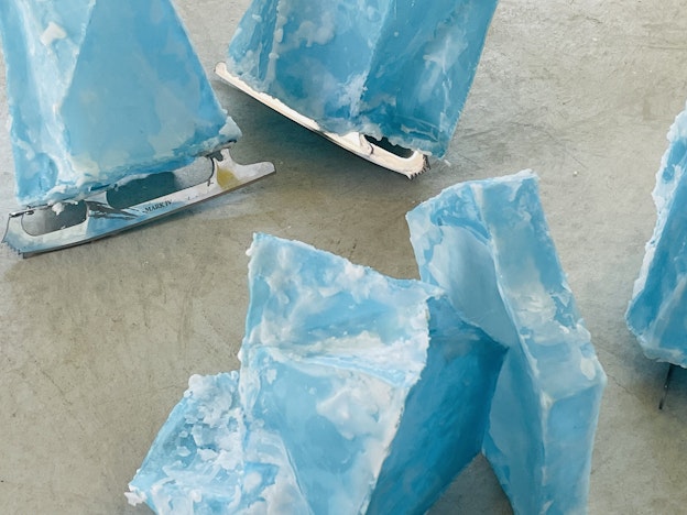 Installation view of “Ice Skating.” 5 ice skates that have shoes made out of blue beeswax,  resembling blocks of ice, are spread throughout a light grey cement gallery floor in varying  angles.