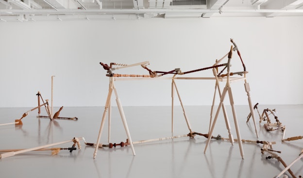 Installation view of “Negotiated Difference,” a rhizomatic sculpture consisting of carved wood  and 3D-printed filaments in wood, metal, and plastic, connected with various objects including  balusters, pipes, and bowling pins. The sculpture spreads throughout the grey floor of a gallery  room, which has white walls and a pipe grid.