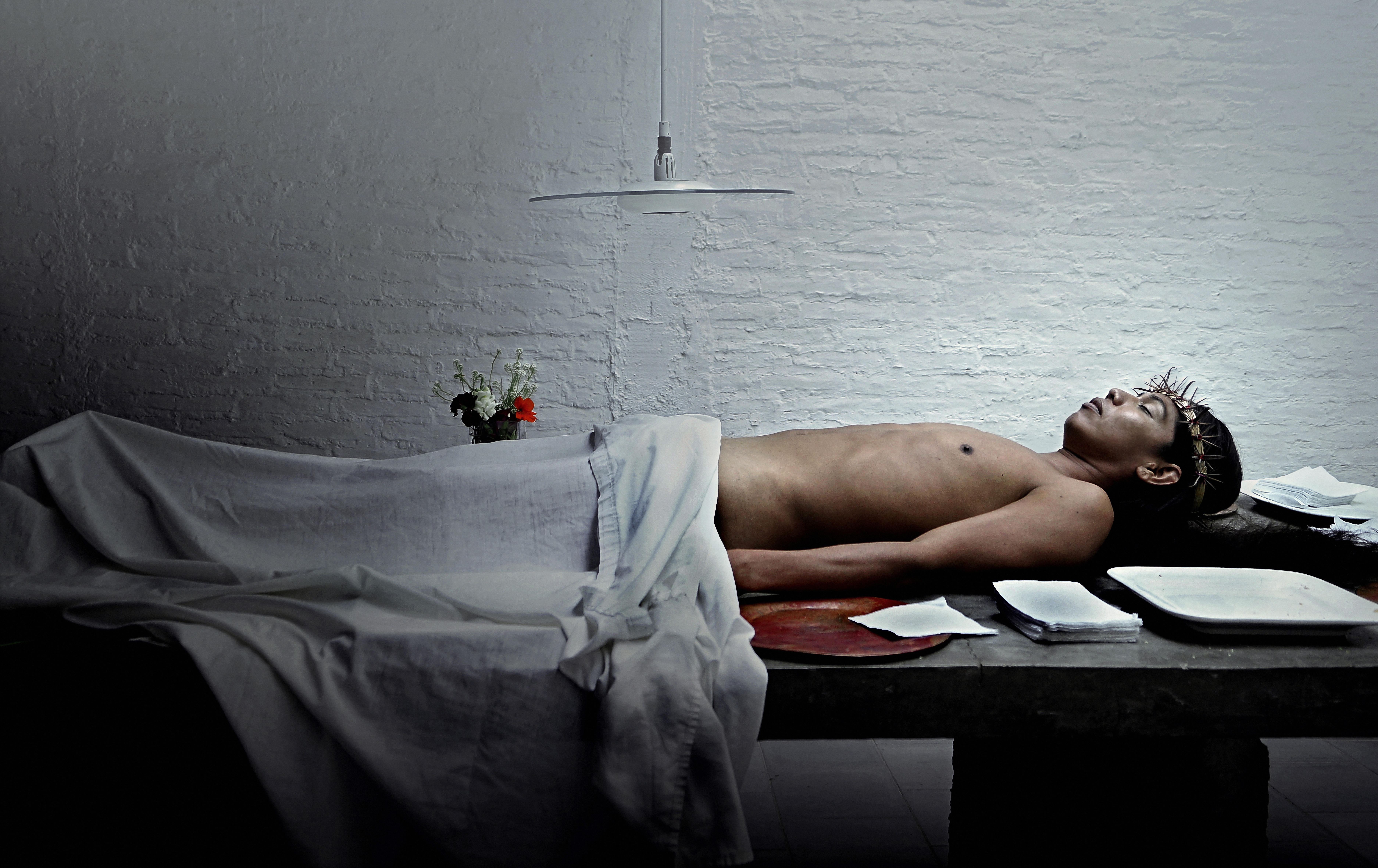 Lukas Avendaño mid-performance, lying down with her back on a stone table, arms to the side and eyes closed. Avendaño is naked, but covered from the waist down by a hospital linen, which matches the bare white brick wall in the background and the white lamp that hangs above the center of her body. White cloths are neatly stacked on the table along her body and head, which is decorated with a gold crown of thorns. There are also several trays and a small bouquet of white, purple, and red flowers beside her.