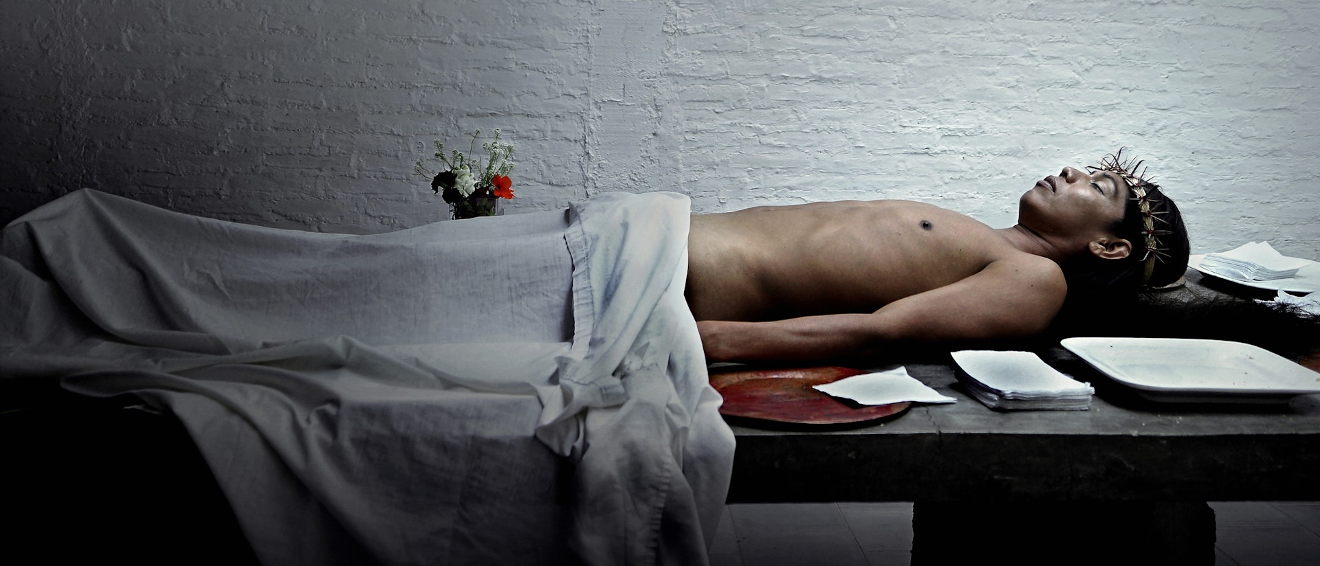 Lukas Avendaño mid-performance, lying down with her back on a stone table, arms to the side and  eyes closed. Avendaño is naked, but covered from the waist down by a hospital linen, which matches  the bare white brick wall in the background and the white lamp that hangs above the center of her body.  White cloths are neatly stacked on the table along her body and head, which is decorated with a gold  crown of thorns. There are also several trays and a small bouquet of white, purple, and red flowers beside her.