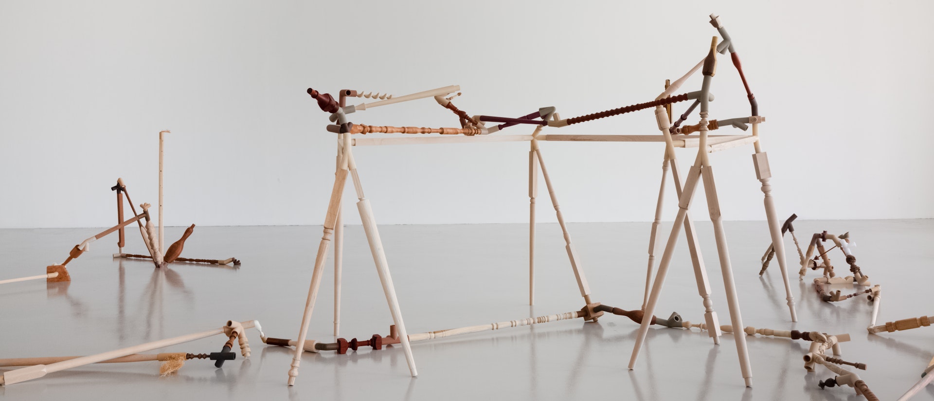 Installation view of “Negotiated Difference,” a rhizomatic sculpture consisting of carved wood  and 3D-printed filaments in wood, metal, and plastic, connected with various objects including  balusters, pipes, and bowling pins. The sculpture spreads throughout the grey floor of a gallery  room, which has white walls and a pipe grid.