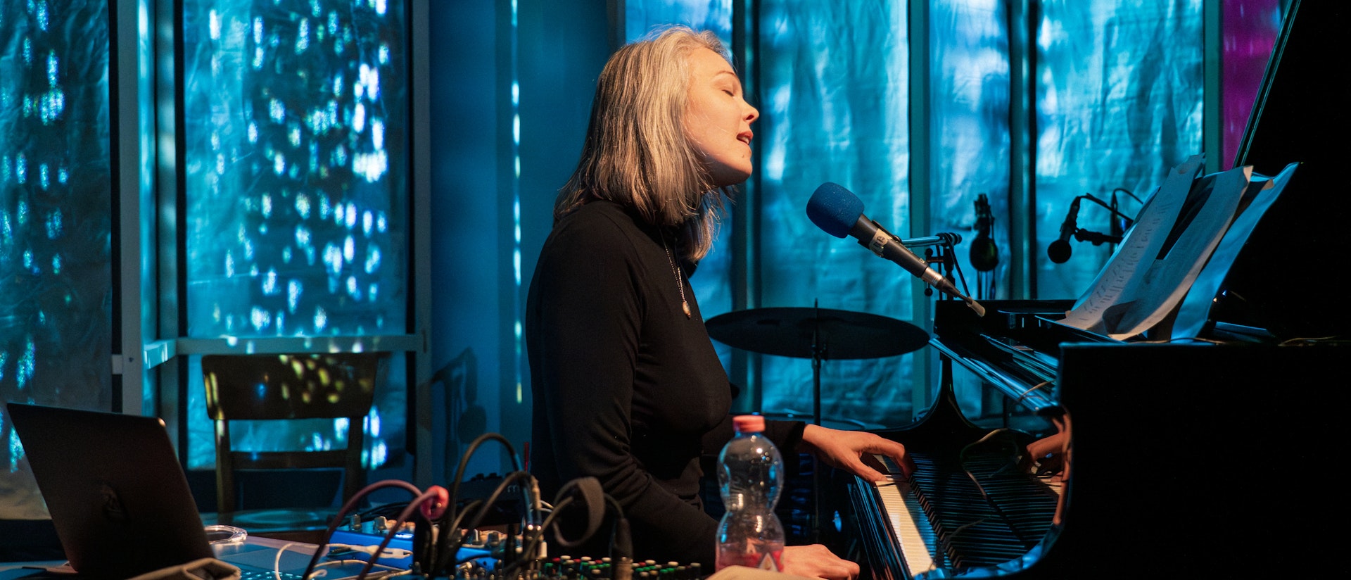 Olivia Block mid-performance. She is sitting down in front of a piano that she is playing with one hand while singing into a microphone that hangs in front of her sheet music. There is a table of electronic equipment with a red beverage in a plastic bottle laid out in front of her. Wires are plugged into and coming out of her gear. Behind Block are room dividers, panels, and a bit of a wall that is lit up with solid blue, sea green, and purple projections, as well as white dots and white amorphous shapes. She is wearing a black turtleneck with a gold necklace over it.