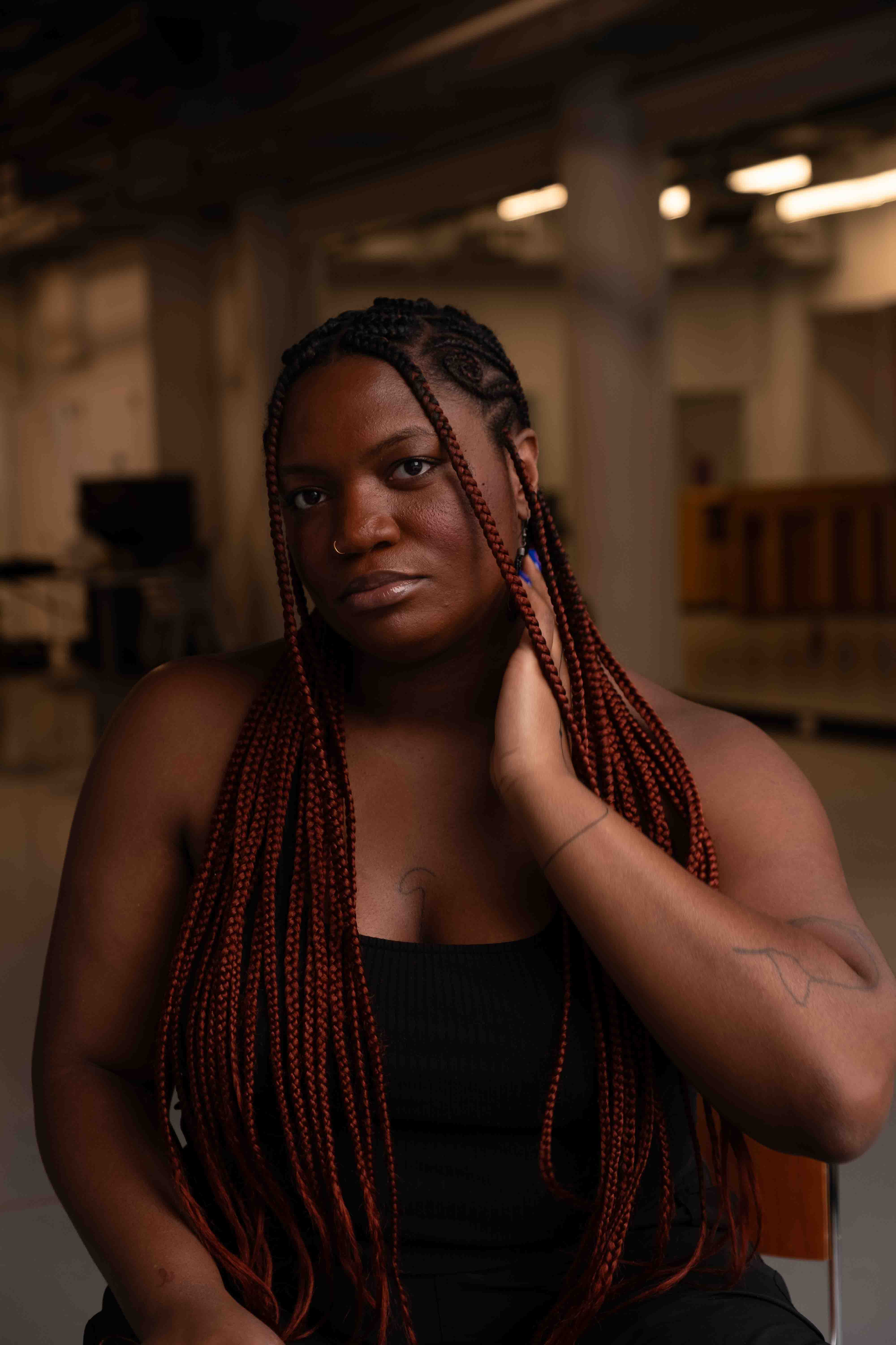 Ogemdi Ude sitting on a wooden and metal chair in a dance studio. She is in front  of the photographer and looking directly at the camera, wearing a blank tank top, black  pants, and with a bright purple manicure. Her left hand is positioned on her neck gently  touching her jawline. Her right hand, partially cut off in the frame, rests on her knee.