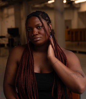 Ogemdi Ude sitting on a wooden and metal chair in a dance studio. She is in front  of the photographer and looking directly at the camera, wearing a blank tank top, black  pants, and with a bright purple manicure. Her left hand is positioned on her neck gently  touching her jawline. Her right hand, partially cut off in the frame, rests on her knee.