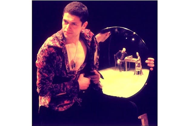 A performer stands in a dark space. They wear an unbuttoned shirt colored with splotches of red and purple and a long pendant. They hold a circular mirror out to the side which reflects the image of two people sitting at a small wooden table. This performer looks intently ahead in the direction the mirror is angled towards. 
