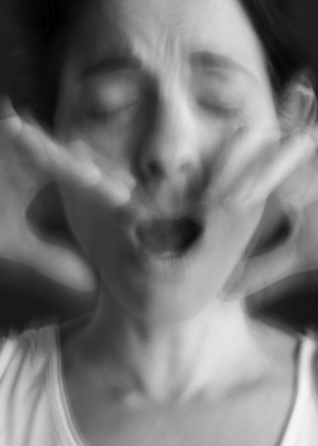 A black and white closeup photograph of vocalist Inbal Hever. The image is blurred as though captured mid-movement, Hever's eyes are closed and her mouth open. Her hands are raised on either side of her mouth, fingers gently curved and framing her jaw. She is wearing a white sleeveless top. 