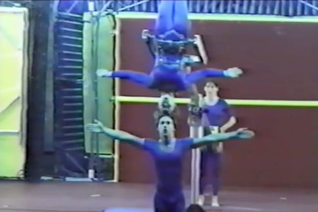 Two gymnasts clad in blue performing. One of them stands with his feet put together and with their arms they are supporting the other gymnast who has the same stance in an upside down manner.