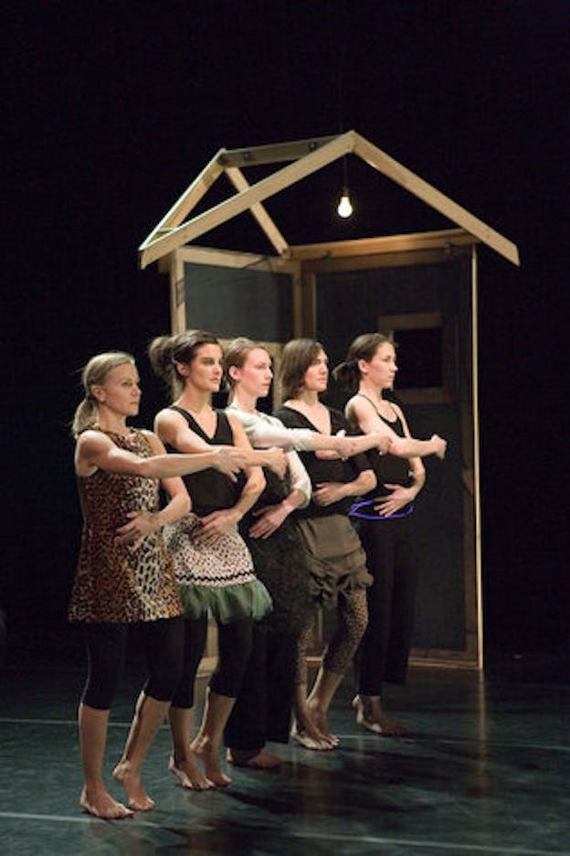 Five performers dressed in mismatched outfits are lined up diagonally facing right in front of a simplified wooden house structure. A light bulb is hung from above to illuminate the interior of the house structure. The performance is staged on a black floor against a black backdrop.