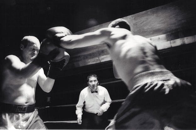 A blurred, black and white image of two people boxing each other, wearing boxing gloves and shorts. In between them, in the background, a person in a white shirt and a bow tie is visible and closely watches the boxers. The background is otherwise bare and dark with one large wooden beam and nothing else. 