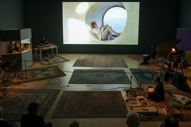 A projection of a figure sitting in cylinder gap in a wall, is surrounded by carpets of flowery design and people sitting on around them with various musical instruments. Cables cross the middle of the room.