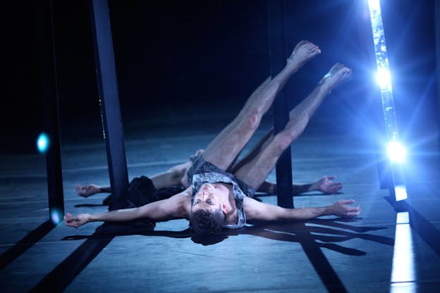 A person laying with their back on the floor and their legs upwards supported by the mirror behind them, lean to the side. Blue and white lights illuminate them.