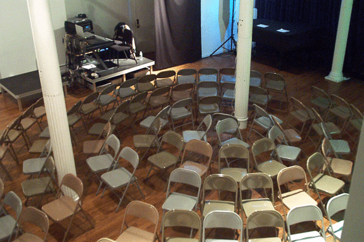 An image of grey folding chairs arranged in a circle around two white columns. Behind this, there is sound equipment and two black folding chairs on an elevated black panel.  