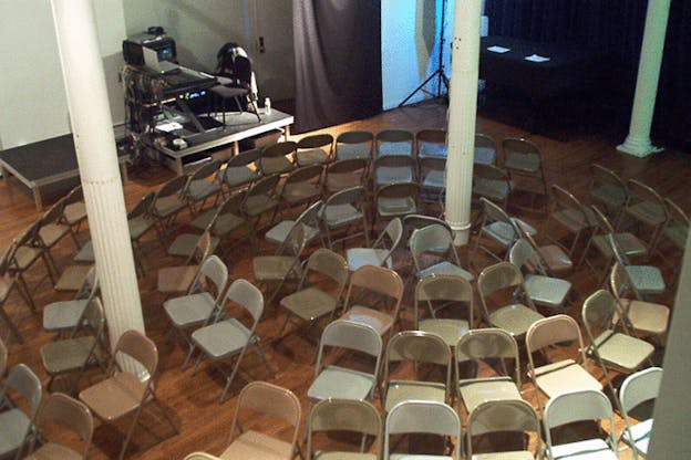 An image of grey folding chairs arranged in a circle around two white columns. Behind this, there is sound equipment and two black folding chairs on an elevated black panel.  