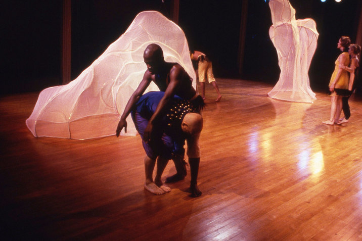 Image of perfromers on stage. Two figures in the front closest to the camera hunch together. One figure hunched their head towards their knees while the other with their arms extended forward hunch on top of the bent figures back.