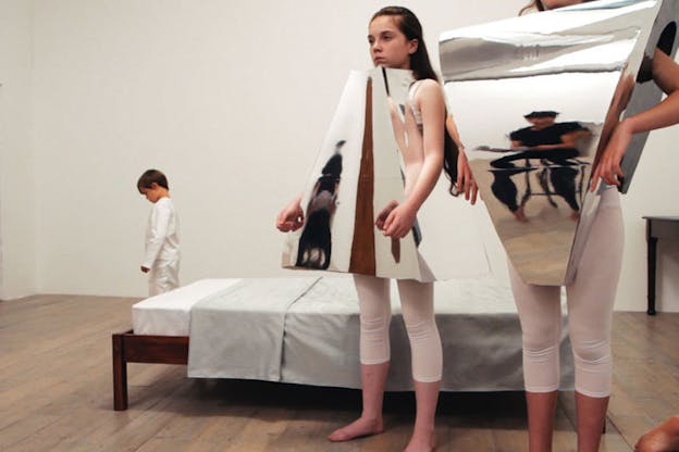 Low-angle shot of two children standing in front of a bed dressed in white leggings and 3D triangular mirror formations around their chest that reflect the image of a seated person dressed in black. In the left corner a young child dressed all in white looks down at the wood floor.