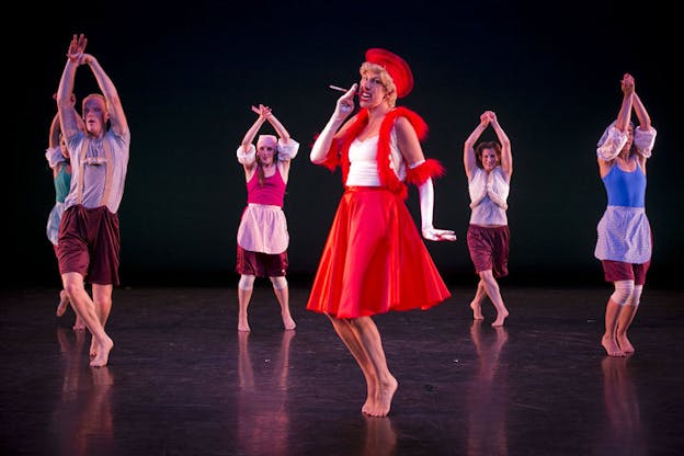 Dancers with their hands upward and linked together are situated behind a person dressed in red, who is smoking a cigarette and stands on their tippy toes. 