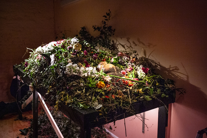 Image still of a person laying down on a big table. Their body is engulfed and hidden by a variety of colorful leaves and flowers. Underneath the table  dead black leaves have been scattered.