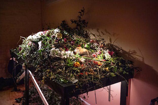 Image still of a person laying down on a big table. Their body is engulfed and hidden by a variety of colorful leaves and flowers. Underneath the table  dead black leaves have been scattered.