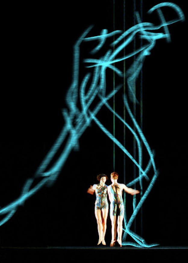 Two performers stand in the lower left of a large dark space. They are on their tip toes and face each other. They wear leotards with a metallic blue toned abstract pattern on them. Above them, a large sketch of a human figure in blue is projected onto an otherwise black background. This human figure appears to be lunging to the right. 