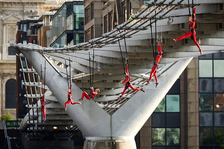 Performers clad in red hang from the edges of a bridge by supportive mechanisms surrounding their waists, in a line formation.