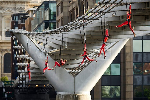Performers clad in red hang from the edges of a bridge by supportive mechanisms surrounding their waists, in a line formation.
