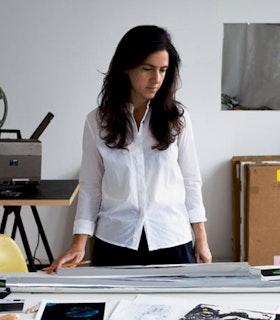 A portrait of Amie Siegel, dressed in a white collared shirt with long brown hair. The artist looking downwards stands in front of a desk with multiple artist materials.