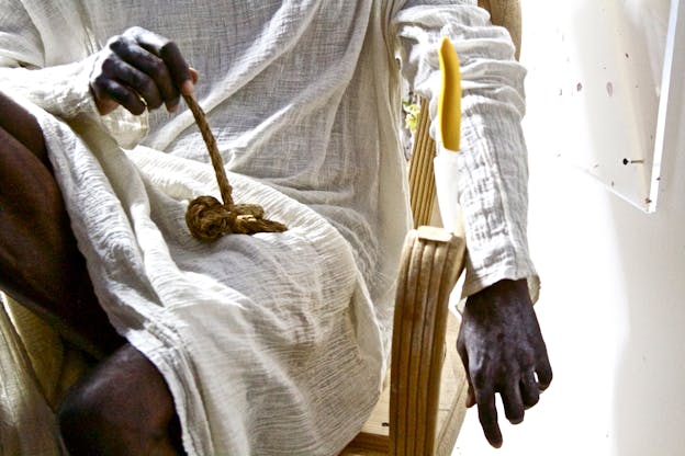 In a performance, David Thomson sits in a wooden chair, wearing a white tunic. He holds a thick knotted string in their lap with one hand and the other extends over the arm of the chair. The wall is white and has a plastic slab nailed into it behind him.