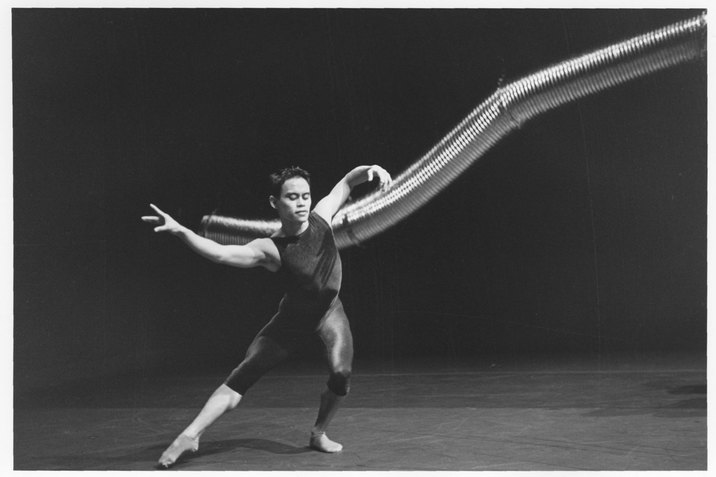 A black and white photograph of a performer dressed in dark rompers in front of a long and curved aluminum duct hung from above. The performer bends their left arm and leg while extending their right arm and leg. The stage has a light color floor and a dark backdrop.