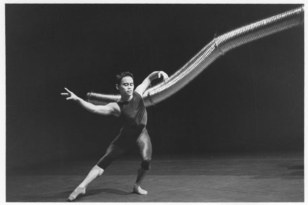 A black and white photograph of a performer dressed in dark rompers in front of a long and curved aluminum duct hung from above. The performer bends their left arm and leg while extending their right arm and leg. The stage has a light color floor and a dark backdrop.