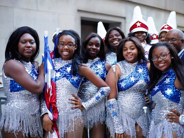 Young people in shiny silver and blue star-decorated color guard uniforms hold flags and batons, smiling in a row.