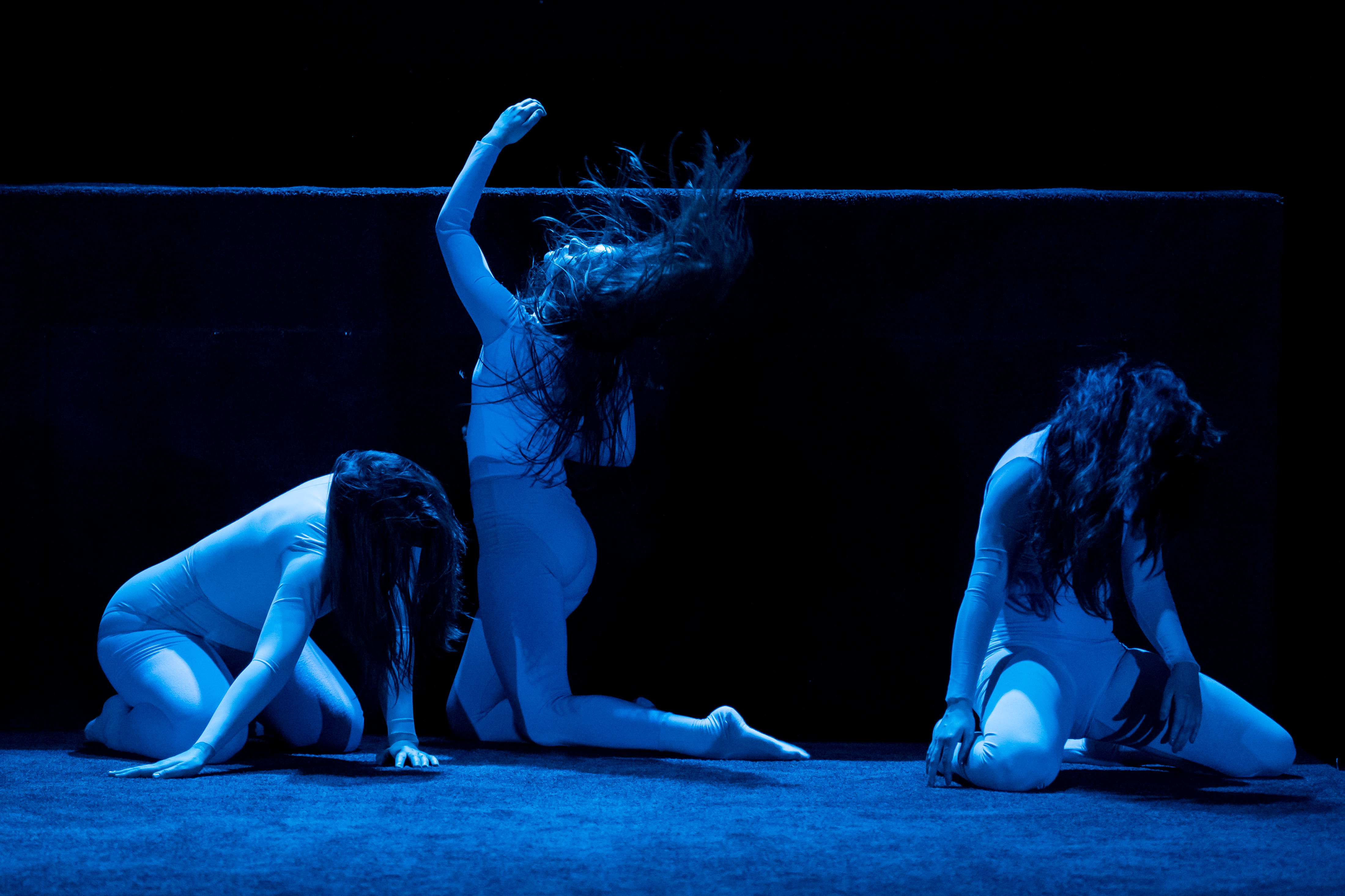 Three performers lighted by blue lights lean on a stage with their heads turned downwards their long hair covering their faces, except the middle one who kneels on the opposite direction her body and hair flung forward and and the left arm turned upwards. 