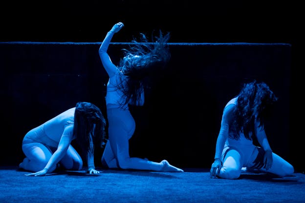 Three performers lighted by blue lights lean on a stage with their heads turned downwards their long hair covering their faces, except the middle one who kneels on the opposite direction her body and hair flung forward and and the left arm turned upwards. 