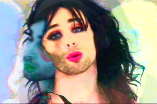 A performer wear bright pink lipstick, heavy eye makeup, and a black wig puckers their lips. In the background, their face is depicted at an angle, without makeup, and hued in blue. Outlines of themselves in various hues are repeated throughout the image. 