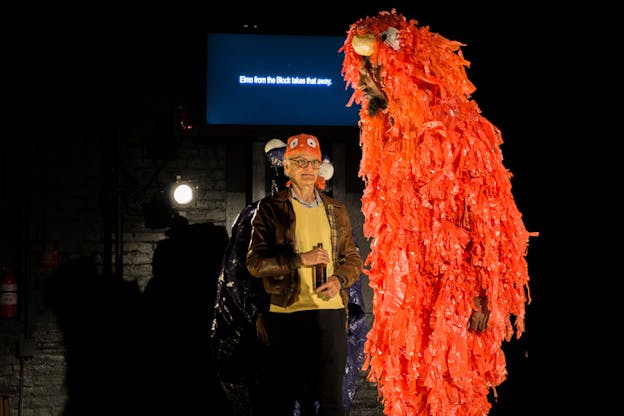 A person holding a mic stands on stage next to a figure wearing an Elmo character costume made of orange crepe paper. Behind them on a small screen are typed the words 