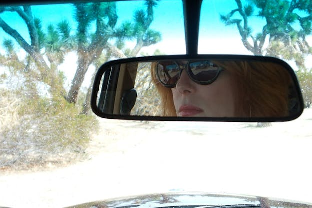 Close-up of a person wearing sunglasses and lipstick's reflection in a rearview mirror, and through the car window, cacti and shrubs are visible.