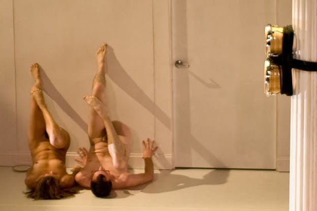 Two people lie naked on the floor, their bottom halfs face upwards with their legs supported by the wall. For each the right foot lies on top of the left legs knee.