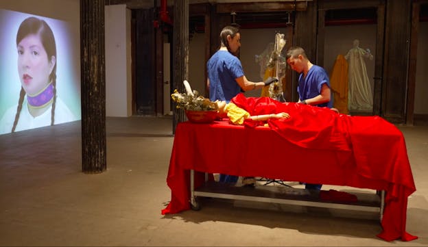 devynn emory and Joseph Pierce stand behind a metal table draped in a red tablecloth. They are dressed in blue scrubs and are wearing black latex gloves. A yellow-clad medical mannequin lies on the table, a bowl of dried flowers next to its head. Elisa Harkins is projected onto a wall to their left.